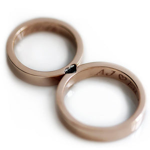 Heart design couples band set by Cadi in 18k rose gold, one ring with diamond, one with black stone; heart and initials inside.