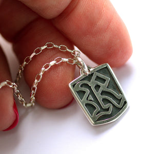 Silver gothic initial letter tag pendant with intricate design held between fingers on a white background