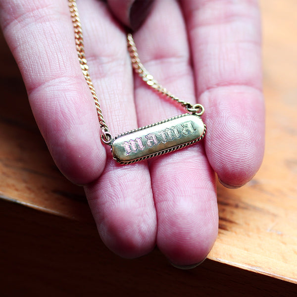 Gold tag necklace with  "mama" ingraved gothic leeters pendant held in a hand 