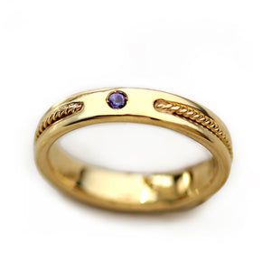 Women’s 18k yellow gold wedding band worn on hand featuring purple sapphire accent stone and handcrafted rope filigree detailing