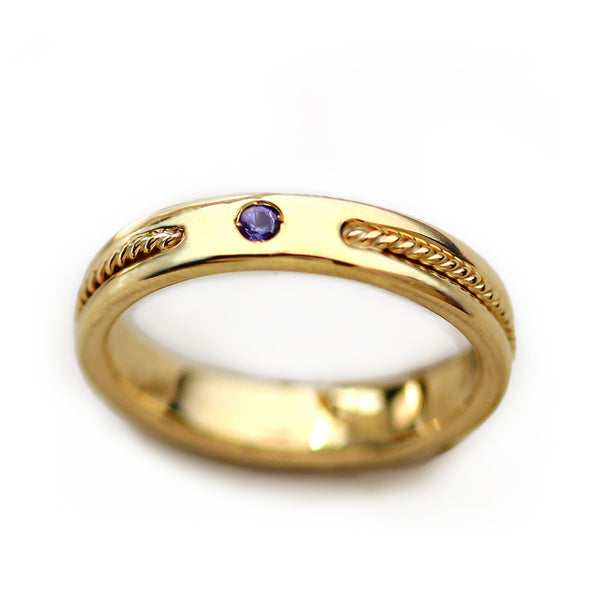 Women’s 18k yellow gold wedding band worn on hand featuring purple sapphire accent stone and handcrafted rope filigree detailing
